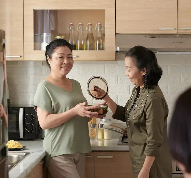 Two women in a kitchen preparing food, with a third person partially visible at the table.