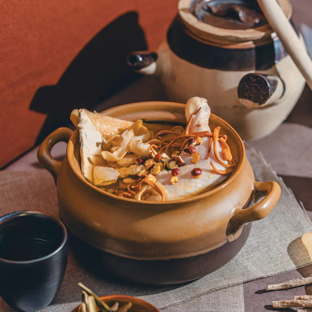 Ceramic pot with confinement soup on a table with a teapot and cup in the background.