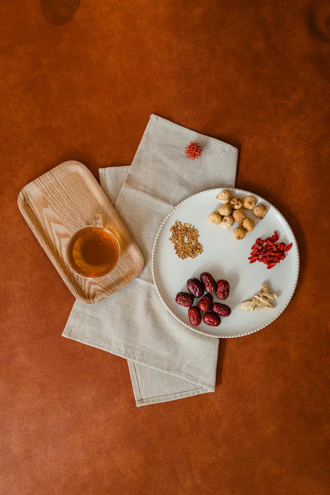 Assorted dried herbs and nuts on a white plate with a wooden tray and napkins on a brown surface.